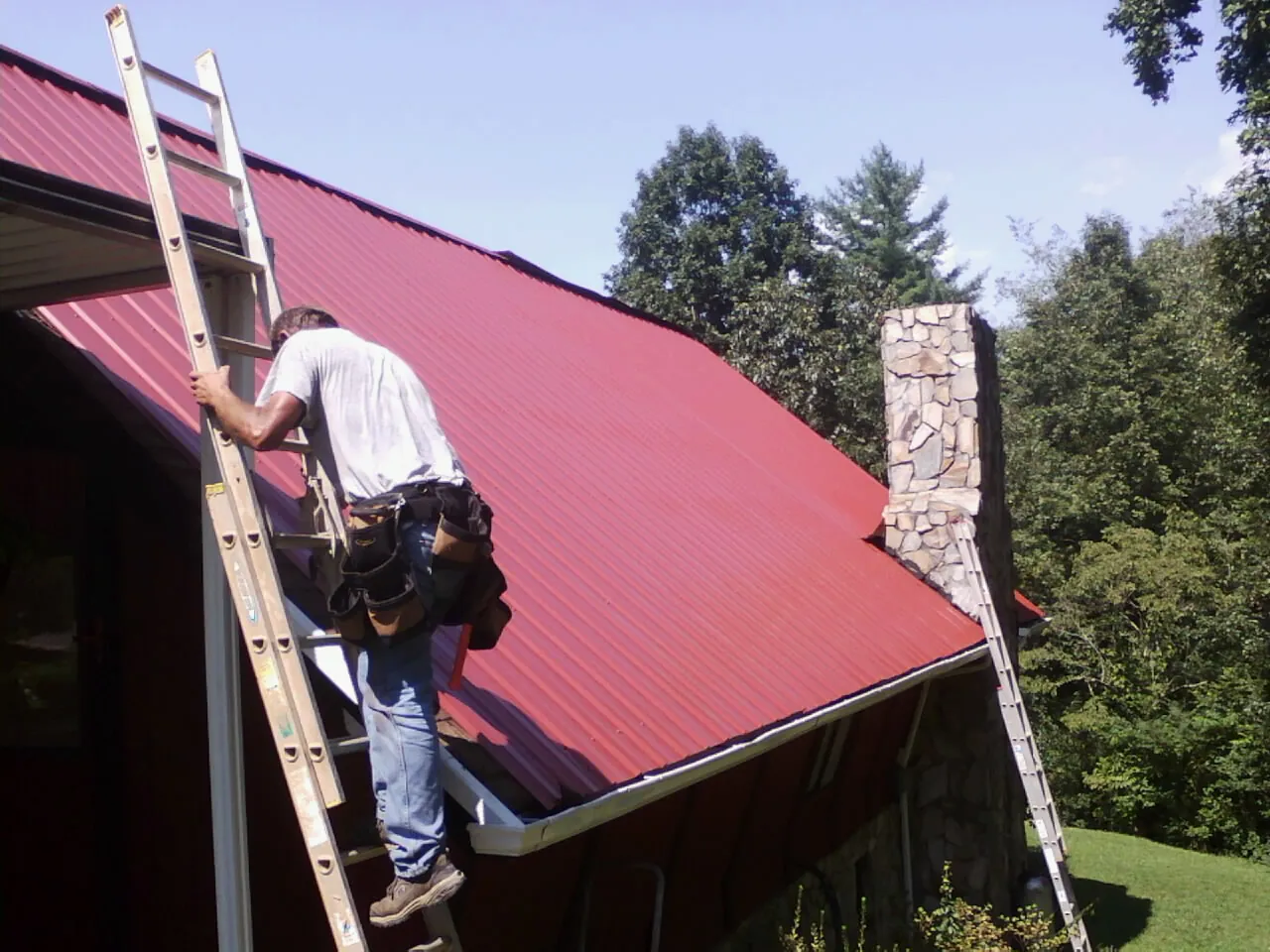 Roofer on a ladder beside a red metal roof and stone chimney — Soffit & Fascia Repair by Roofing Brothers in Vacaville